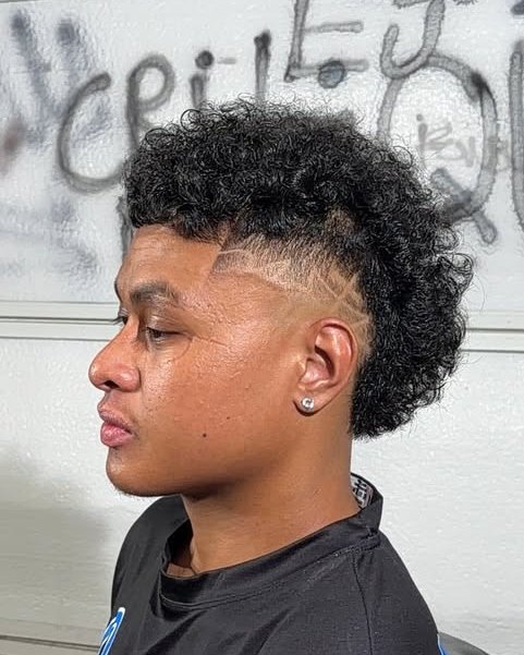 Curly burst fade mohawk with a razor design line, side profile against the CRITIQUE wall